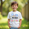 Child wearing a white t-shirt with colorful designs and text outdoors