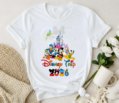 White t-shirt with Disney-themed design featuring characters and 'Disney Trip' text, placed on a light surface with flowers and a mug.
