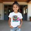 Young girl wearing a white t-shirt with colorful text and graphics, standing outdoors.