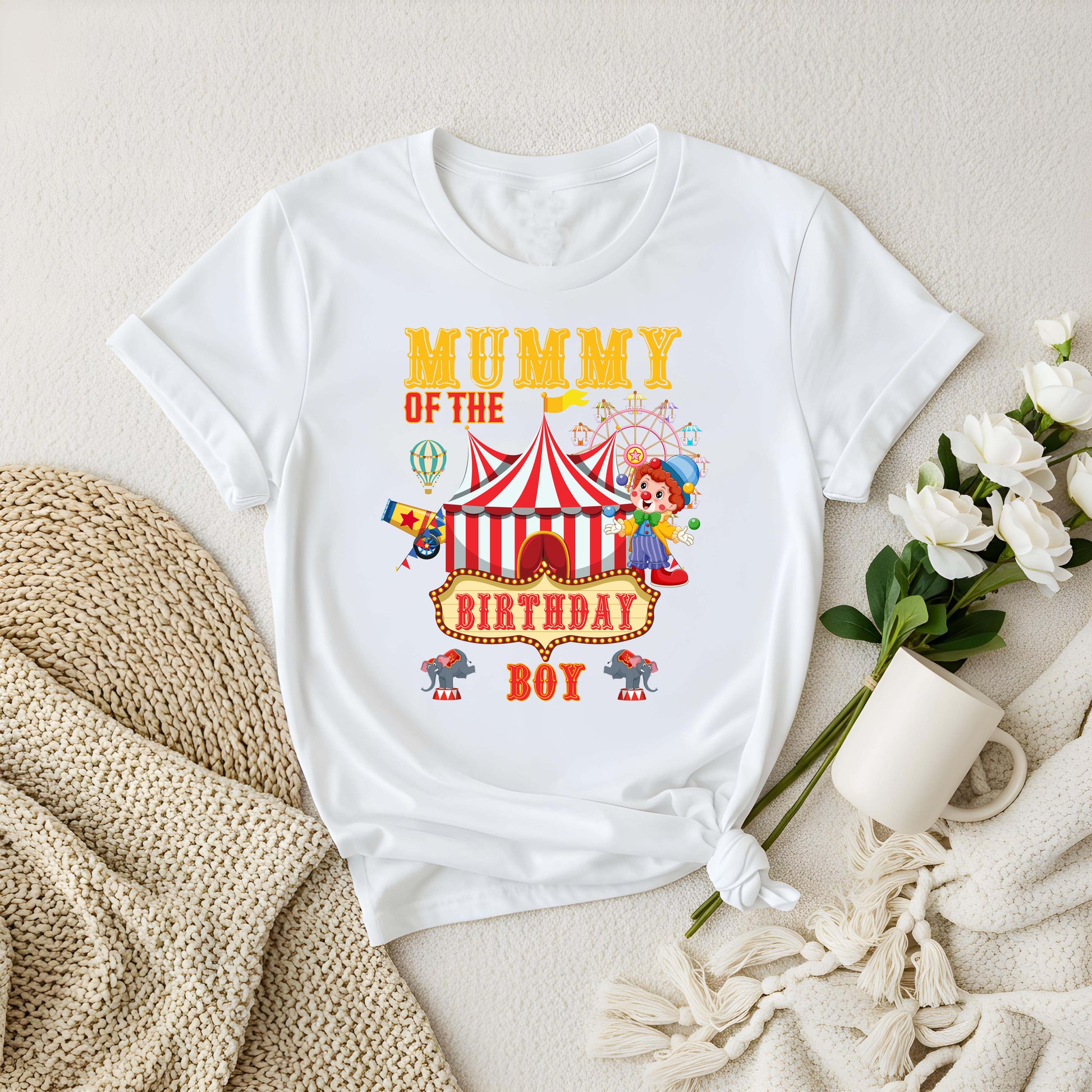 White t-shirt with circus-themed text on a beige surface with flowers and a mug.