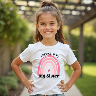 Young girl wearing a 'Promoted to Big Sister' t-shirt with a rainbow design outdoors.