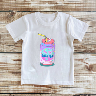 White t-shirt with a colorful 'Little Soda Pop' design on a wooden surface