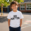 A young boy smiling in an outdoor schoolyard, wearing a white short-sleeve t-shirt with the text 'YOU MATTER' printed in black across the chest, paired with navy blue pants.