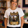 A smiling woman wearing a white graphic T-shirt with a colorful wizard-themed 'Book Week Quest Ms Grater Wizard Academy' print, standing in a blurred library or classroom setting.
